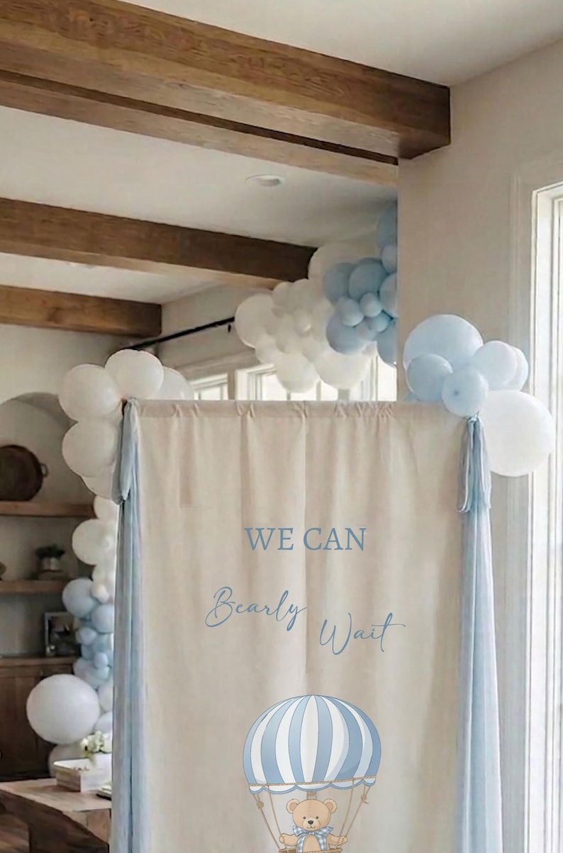 May include: A rectangular beige fabric backdrop featuring the blue text WE CAN Bearly Wait and a graphic of a teddy bear in a blue and white striped hot air balloon. The backdrop is framed by light blue fabric panels and white and blue balloons.
