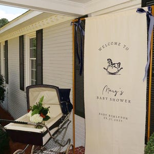 May include: A vintage baby carriage with a white interior and a bouquet of flowers sits next to a banner that reads "Welcome to Mary's Baby Shower." The banner has a rocking horse graphic and the date 09.21.2025.