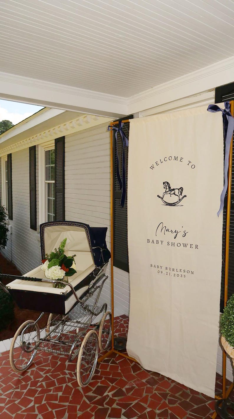 May include: A vintage baby carriage with a white interior and a bouquet of flowers sits next to a banner that reads "Welcome to Mary's Baby Shower." The banner has a rocking horse graphic and the date 09.21.2025.