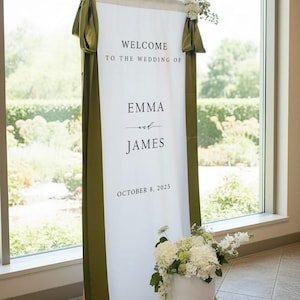 K&ouml;nnte beinhalten: Ein hohes, vertikales Hochzeits-Willkommensschild in Wei&szlig; mit dem schwarzen Text "WELCOME TO THE WEDDING OF EMMA and JAMES OCTOBER 8, 2025". Das Stoffbanner ist mit olivgr&uuml;nen B&auml;ndern drapiert und mit wei&szlig;en Blumenarrangements verziert.