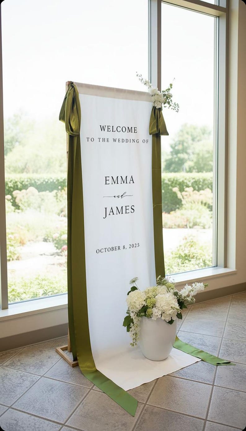 K&ouml;nnte beinhalten: Ein hohes, vertikales Hochzeits-Willkommensschild in Wei&szlig; mit dem schwarzen Text "WELCOME TO THE WEDDING OF EMMA and JAMES OCTOBER 8, 2025". Das Stoffbanner ist mit olivgr&uuml;nen B&auml;ndern drapiert und mit wei&szlig;en Blumenarrangements verziert.