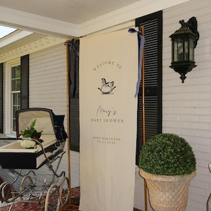 May include: A baby shower welcome sign with the text "Welcome to Mary's Baby Shower" and a rocking horse graphic. The sign is cream-colored and framed with wooden supports. A vintage baby carriage is in the foreground.