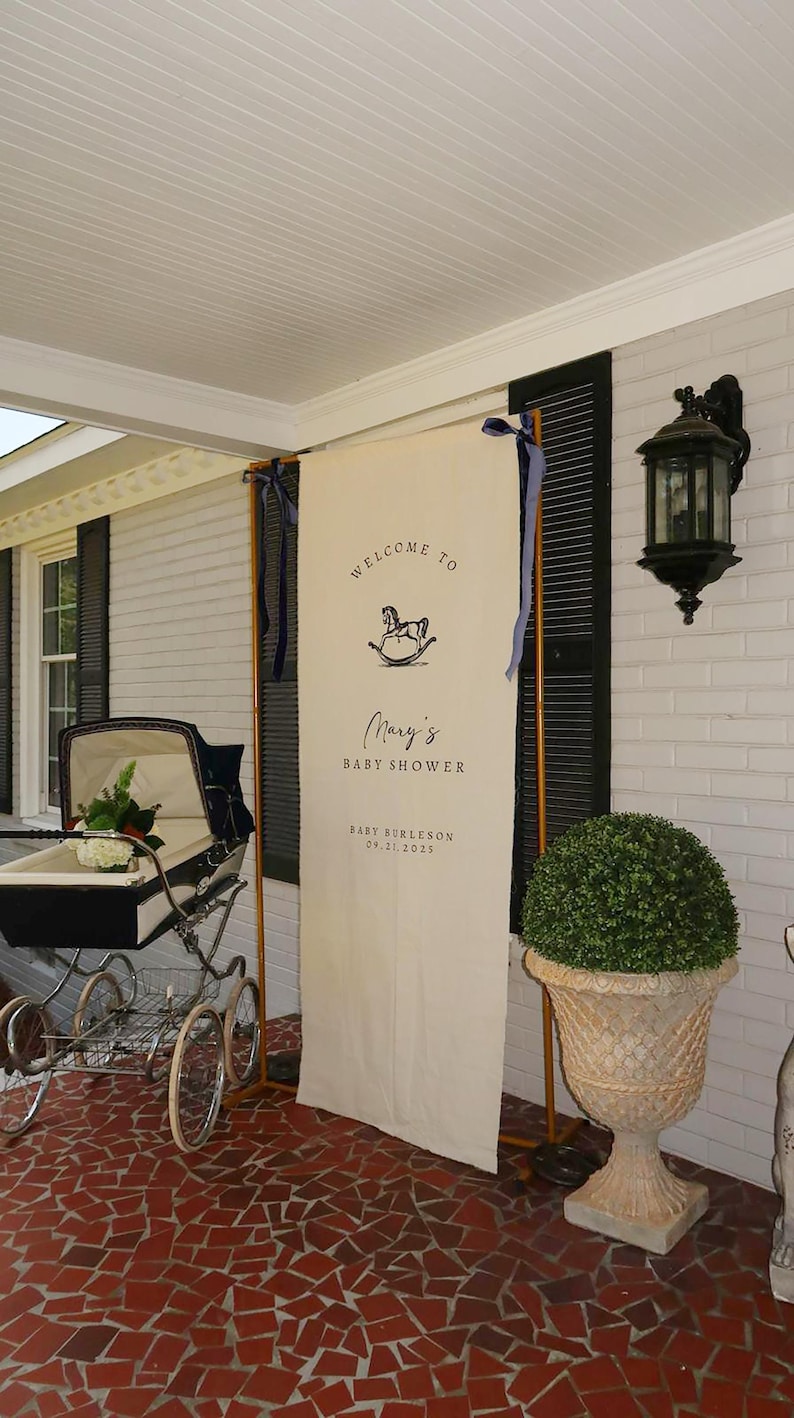 May include: A baby shower welcome sign with the text "Welcome to Mary's Baby Shower" and a rocking horse graphic. The sign is cream-colored and framed with wooden supports. A vintage baby carriage is in the foreground.
