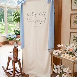 Pode incluir: Uma faixa alta e retangular de tecido off-white suspensa em uma estrutura de metal dourado, com o texto impresso em preto "for this child I have prayed 1 SAMUEL 1:27". A faixa é decorada com fitas de cetim azul claro nos cantos superiores.