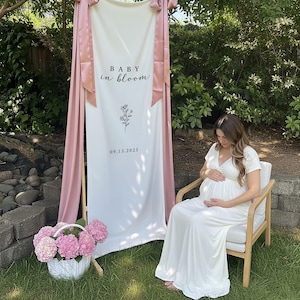 May include: A white banner with the text "BABY in bloom" and the date 09.13.2025, decorated with a floral illustration. Pink fabric drapes on either side. A white basket of pink flowers sits on the grass. A person in a white dress sits in a wooden chair.