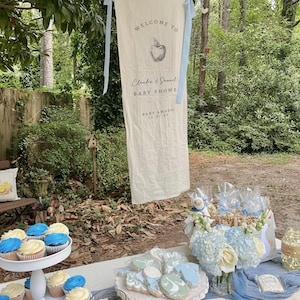 Może przedstawiać: Kremowy baner na baby shower z napisem "WELCOME TO" i "Claudia & Samuel BABY SHOWE" z grafiką jabłka. Babeczki i ciasteczka są wystawione na zewnątrz.
