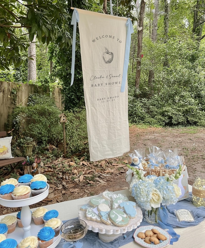 Może przedstawiać: Kremowy baner na baby shower z napisem "WELCOME TO" i "Claudia & Samuel BABY SHOWE" z grafiką jabłka. Babeczki i ciasteczka są wystawione na zewnątrz.