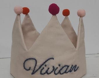 Op de afbeelding: Een witte stoffen kroon met een blauwe geborduurde naam "Vivian" en roze, oranje en rode pompoenen bovenop.