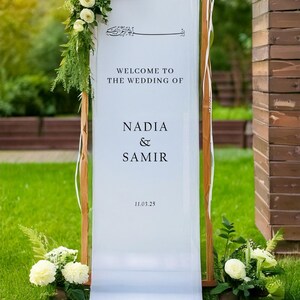 May include: White wedding sign with calligraphy in Arabic script, the text "Welcome to the wedding of Nadia & Samir 11.03.25" and white flowers on either side.