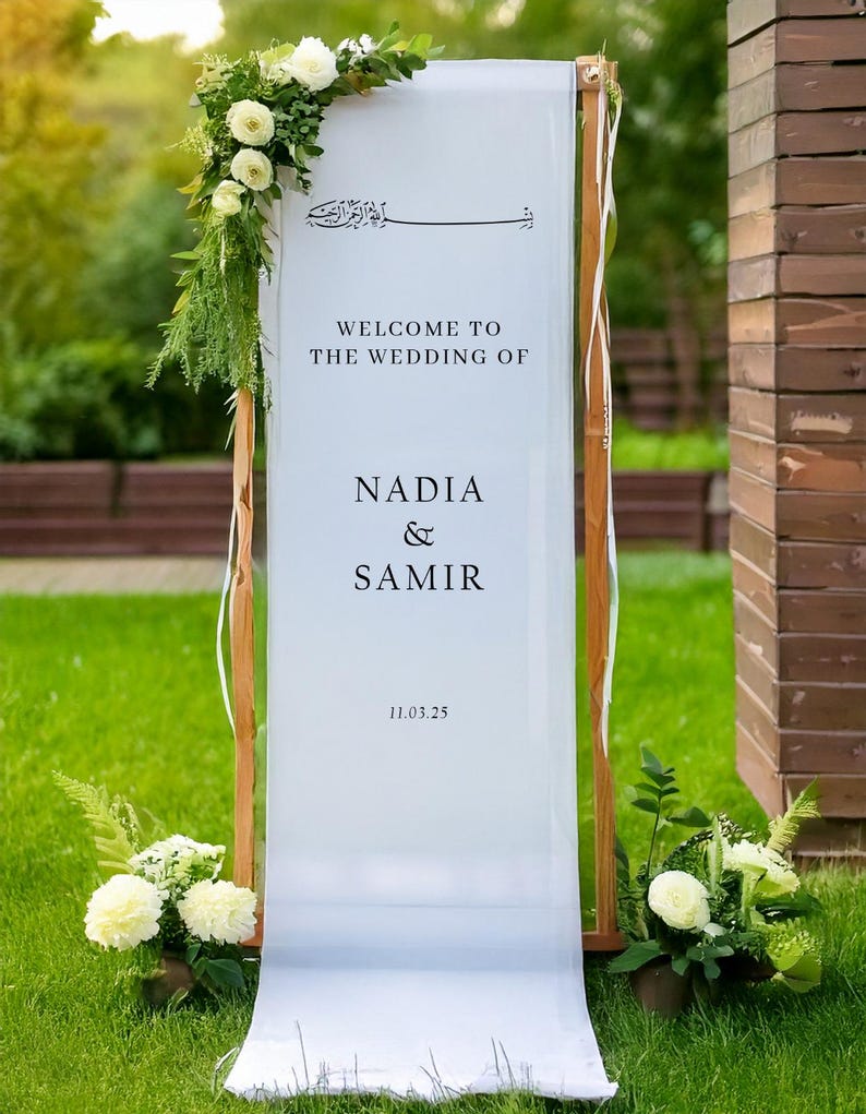 May include: White wedding sign with calligraphy in Arabic script, the text "Welcome to the wedding of Nadia & Samir 11.03.25" and white flowers on either side.