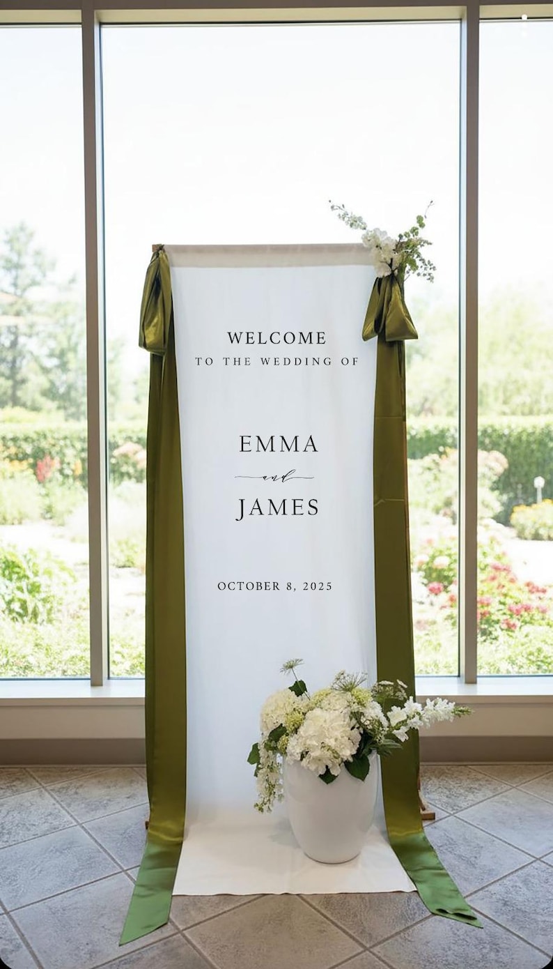 K&ouml;nnte beinhalten: Ein hohes Willkommensschild aus wei&szlig;em Stoff f&uuml;r eine Hochzeit mit dem Text "WELCOME TO THE WEDDING OF EMMA and JAMES OCTOBER 8, 2025". Das Schild ist mit langen olivgr&uuml;nen Satinb&auml;ndern an den Seiten und wei&szlig;en Blumenarrangements verziert.