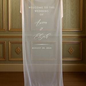 May include: A sheer white wedding welcome sign with the text "WELCOME TO THE WEDDING OF Aurora & Elijah AUGUST 20, 2024". The sign is suspended from a gold rod with pink ribbons.