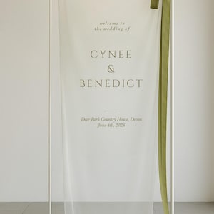 May include: A white wedding welcome sign with the names "CYNEE & BENEDICT" in a soft green font. The sign includes the wedding date and location, with a green ribbon accent. The sign is displayed on a white metal frame.