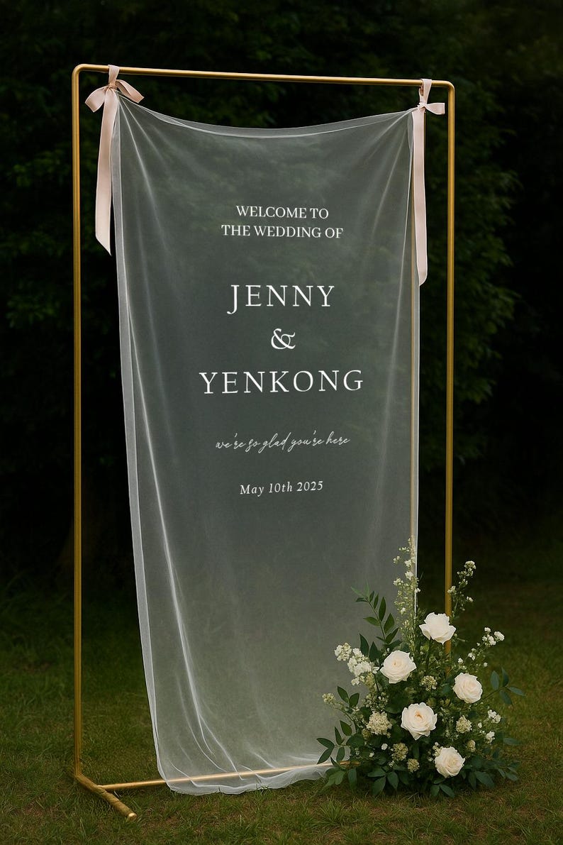 May include: A gold metal wedding sign with a sheer white fabric backdrop. The sign reads "WELCOME TO THE WEDDING OF JENNY & YENKONG" with the date "May 10th 2025". White roses and greenery decorate the base.