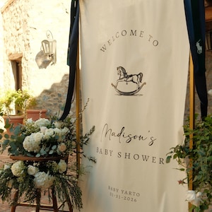 Może przedstawiać: Kremowy baner z napisami "Welcome to" i "Madison's Baby Shower" czarnym pismem. Ilustracja konia na biegunach znajduje się nad imieniem. Data 01-31-2026 jest wydrukowana na dole. Baner jest ozdobiony ciemnoniebieskimi wstążkami i kompozycjami kwiatowymi.