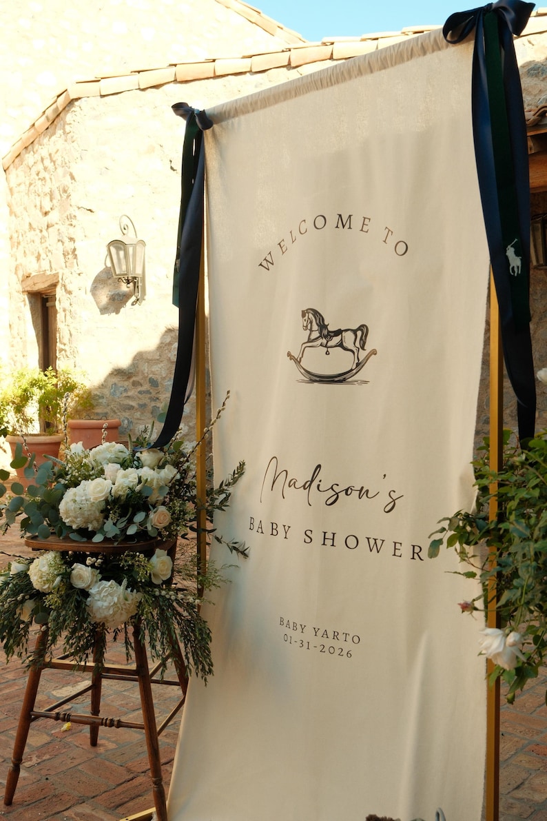 Może przedstawiać: Kremowy baner z napisami "Welcome to" i "Madison's Baby Shower" czarnym pismem. Ilustracja konia na biegunach znajduje się nad imieniem. Data 01-31-2026 jest wydrukowana na dole. Baner jest ozdobiony ciemnoniebieskimi wstążkami i kompozycjami kwiatowymi.