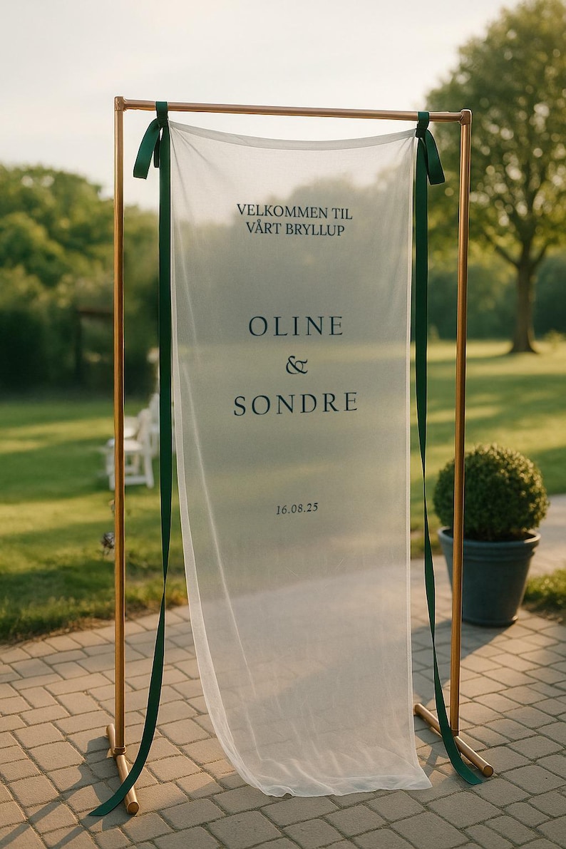 May include: A wedding sign with a sheer white fabric banner suspended from a gold metal frame. The banner has dark green ribbons and features the names "Oline & Sondre" and the date "16.08.25" in black text. The sign is outdoors.