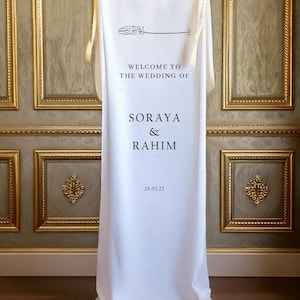 May include: White wedding welcome sign with gold ribbon ties. The sign reads "Welcome to the Wedding of Soraya & Rahim 29.03.25".