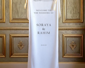 Pode incluir: Uma placa de casamento vertical em tecido branco com texto preto que diz Welcome to the wedding of Soraya & Rahim, com a data 29.03.25 e caligrafia árabe no topo. A faixa é fixada no topo com fitas douradas claras.