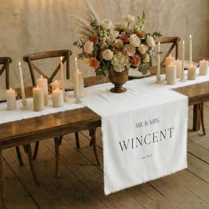 Op de afbeelding: Een houten eettafel gedekt voor een speciale gelegenheid. Een witte tafelloper met "MR. & MRS. WINCENT est. 2023" is gecentreerd. De tafel is versierd met kaarsen, een bloemstuk en couverts.
