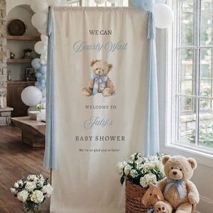 May include: A tall, rectangular cream-colored fabric banner featuring a brown teddy bear illustration with a blue bow, the text "WE CAN Bearly Wait WELCOME TO July's BABY SHOWER We're so glad you're here!", and light blue fabric side drapes.