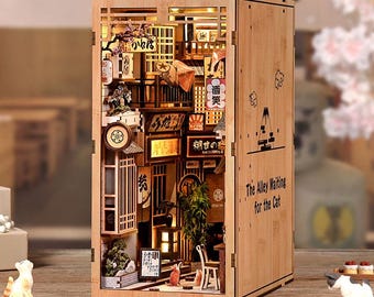 May include: A wooden book nook diorama featuring a miniature Japanese alleyway scene with illuminated lanterns, signs with Japanese text, a small chair, and bamboo plants. The side panel is engraved with the text The Alley Waiting for the Cat.