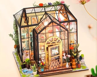 May include: A miniature greenhouse model with glass walls and a wooden frame. The model features a small pond, potted plants, and a variety of flowers. The roof is adorned with greenery and colorful flowers. The door has a small sign.