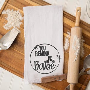 May include: A white tea towel with the text "You Remind Me of the Babe" in a black circular design. The towel is on a wooden chopping board with flour, a rolling pin, and a metal scoop.