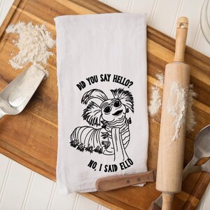 May include: White kitchen towel with a black illustration of a cartoon creature and the text "DID YOU SAY HELLO? NO, I SAID ELLO." The towel is on a wooden cutting board with flour, a rolling pin, and a scoop.