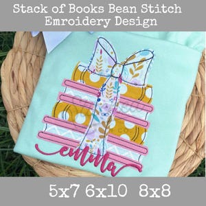 May include: A light blue fabric with an embroidered design of a stack of books tied with a bow. The books are yellow with white polka dots and pink spines. The bow is patterned with floral designs. The name "Emma" is embroidered in pink cursive.