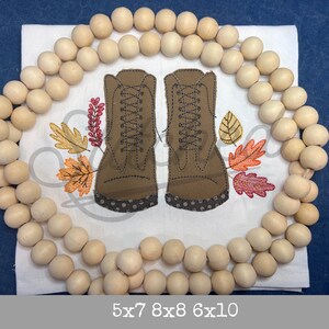 Puede incluir: Diseño bordado de botas marrones con cordones y un patrón de lunares marrones y blancos. Las botas están rodeadas de una corona de cuentas de madera y hojas de otoño en tonos naranja, rojo y amarillo.