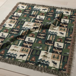 May include: A woven throw blanket with a repeating pattern of outdoor scenes. The design includes moose, fish, canoes, cabins, trees, and loons in a grid of green, cream, and brown squares. The blanket has a fringed edge.