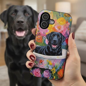 May include: A colorful phone case featuring a black Labrador Retriever in a floral design. The case has a vibrant background with pink, yellow, and orange flowers. A real black Labrador is in the background, out of focus.