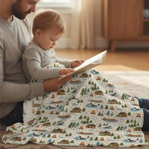 Könnte beinhalten: Eine weiße Decke mit einem Naturmuster mit Hütten, Kanus, Bäumen und Enten. Die Decke ist über einer Person und einem Kind drapiert, die zusammen ein Buch lesen. Die Decke ist weich und gemütlich.
