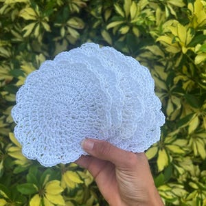 May include: A stack of white, crocheted coasters with a scalloped edge. The coasters are held in a hand, with a background of green and yellow foliage.