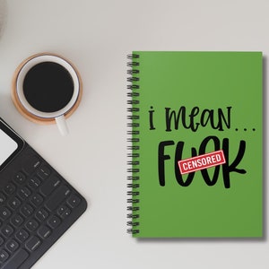 May include: A green spiral-bound notebook with the text "i mean... FUCK" in black, with a red "CENSORED" stamp over the word "FUCK". A cup of coffee and a black keyboard are also visible.