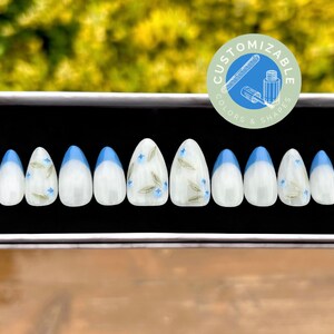May include: A set of ten white oval-shaped press-on nails with a blue tip and a floral design. The nails are in a black box with a white label that says "Customizable Colors & Shapes".