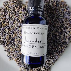 May include: A dark blue glass bottle of "Lavender Baking Extract" with a black cap, set against a backdrop of dried lavender buds. The label reads "Uncommon Extract, Handcrafted, 1 fl oz (30ml)."
