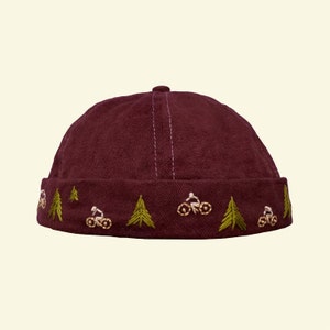 May include: A maroon beanie with an embroidered design of green pine trees and white figures riding bicycles.