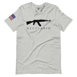 Peut inclure: T-shirt gris clair avec un dessin noir d'un fusil et le mot "NECESARIO" imprimé en dessous. Le drapeau américain est sur la manche gauche. Le t-shirt a un col rond et des manches courtes.