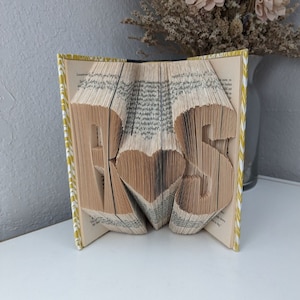 May include: A book art sculpture with the letters "RS" and a heart in the center. The book is folded into a three-dimensional shape and has a yellow and white patterned cover.