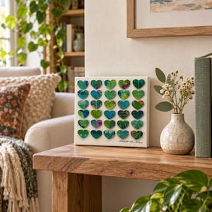 May include: A square white canvas featuring a grid of thirty small, raised heart-shaped cutouts. Each heart displays a unique abstract pattern in shades of green, blue, and magenta. The artist's signature and the year 2024 are written in black ink.