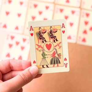 Puede incluir: Un naipe de As de Corazones con un corazón rojo central rodeado por dos músicos tocando el violín y dos figuras bailando. La carta tiene símbolos 'A' rojos en las esquinas sobre un fondo color crema con ilustraciones detalladas.