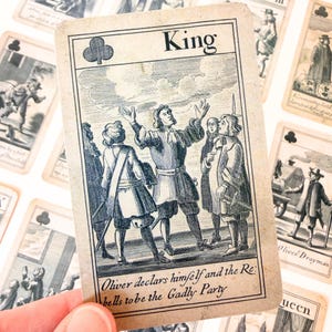 Puede incluir: Carta de juego antigua con una ilustración en blanco y negro de un rey y otras figuras. La carta muestra la palabra "King" y un símbolo de trébol. El texto en la parte inferior dice: "Oliver se declara a sí mismo y a los rebeldes como el partido Gadly." Otras cartas son visibles en el fondo.