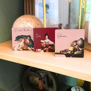 May include: Three square coasters featuring classical art prints. The left coaster shows figures with the text 'Paix & Abondance', the centre is deep red with a portrait and 'Fierté', and the right is light pink with a skull and 'Éphémère'.