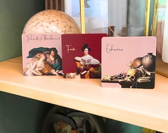 May include: Three square coasters featuring classical art prints. The left coaster shows figures with the text 'Paix & Abondance', the center is deep red with a portrait and 'Fierté', and the right is light pink with a skull and 'Éphémère'.
