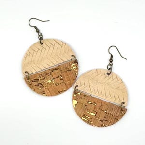 May include: A pair of round earrings. Each earring is divided diagonally, with a textured beige top and a cork bottom with gold flecks. The earrings have bronze-colored hooks and connecting rings.
