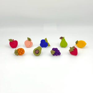 May include: A collection of colorful fruit-shaped decorative items, including a red apple, peach, blueberries, green pear, yellow lemon, pineapple, avocado, purple grapes, and a red strawberry. Each piece has a detailed design and a small green leaf.