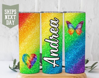 Könnte beinhalten: Drei Regenbogen-Glitzerbecher mit verschiedenen Designs. Der erste Becher hat ein Regenbogenherz, der zweite Becher hat den Namen "Andrea" und der dritte Becher hat einen Regenbogen-Schmetterling.
