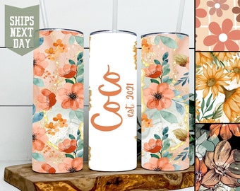 Könnte beinhalten: Drei Edelstahlbecher mit Blumenmustern. Der Becher links hat ein rosa-orangenes Blumenmuster mit goldenen Akzenten. Der Becher in der Mitte ist weiß mit dem Text "Coco est. 2021" in Orange. Der Becher rechts hat ein ähnliches Blumenmuster wie der Becher links.